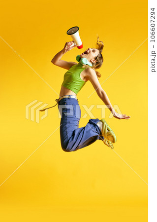 Full-length image of active, positive, young women in stylish clothes jumping with megaphone against yellow studio background. News Full-length image of active, positive, young women in stylish clothes jumping with megaphone against yellow studio background. News 106607294