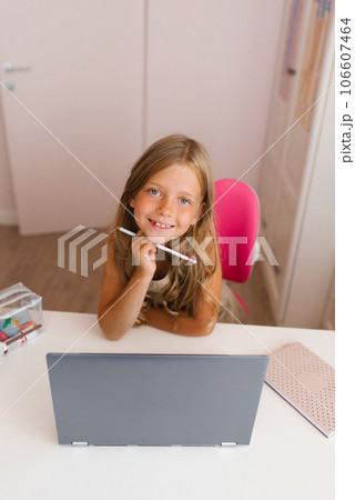 Little girl using a laptop at home for online education and home schooling, looks at the camera with a smile at home and holds a pen in her hand. Distance learning for children 106607464