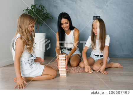 Two children sisters and mom enthusiastically playing a game of building a tower of wooden blocks at home Two children sisters and mom enthusiastically playing a game of building a tower of wooden blocks at home 106607499