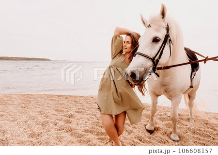 A white horse and a woman in a dress stand on a beach, with the sky and sea creating a picturesque backdrop for the scene. 106608157
