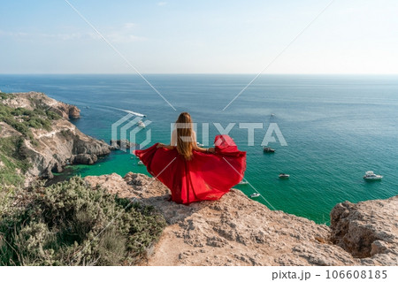 Woman sea red dress yachts. A beautiful woman in a red dress poses on a cliff overlooking the sea on a sunny day. Boats and yachts dot the background. Woman sea red dress yachts. A beautiful woman in a red dress poses on a cliff overlooking the sea on a sunny day. Boats and yachts dot the background. 106608185