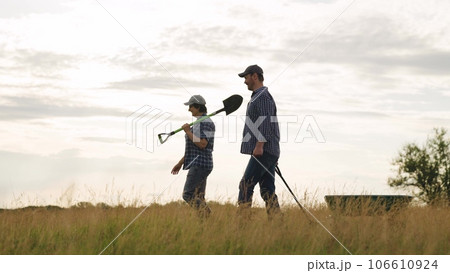Positive farmers family with gardening tools walks along agricultural field after work day. Farm workers man and woman with tools during harvesting at dusk country. Farmers walk along sunset meadow 106610924