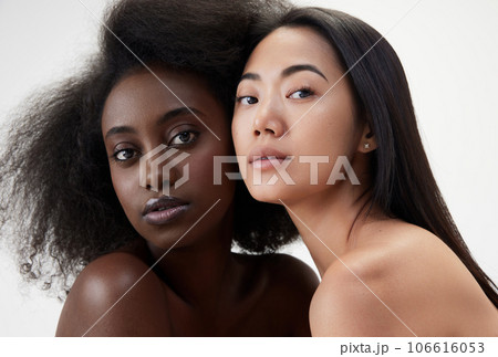 Two beautiful young women, african and asian with well-kept skin, looking at camera against white background Two beautiful young women, african and asian with well-kept skin, looking at camera against white background 106616053
