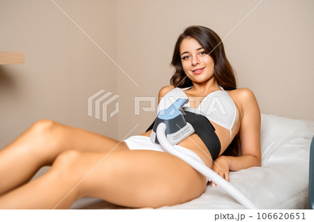 A young and joyful brunette reclines on a beauty salons couch, undergoing a body sculpting procedure with a radiant smile.  106620651
