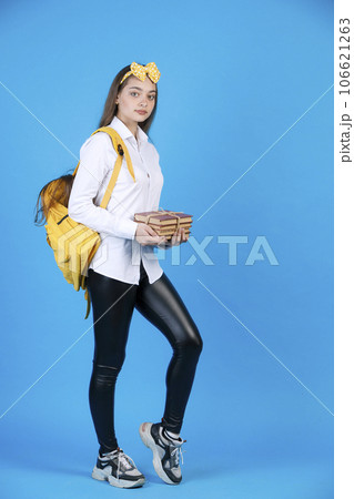 Nice female student in lovely headband carrying bunch of textbooks, while looking at camera. Side view of calm girl in shirt standing, shyly bending one leg, isolated on blue. Concept of studying. Nice female student in lovely headband carrying bunch of textbooks, while looking at camera. Side view of calm girl in shirt standing, shyly bending one leg, isolated on blue. Concept of studying. 106621263