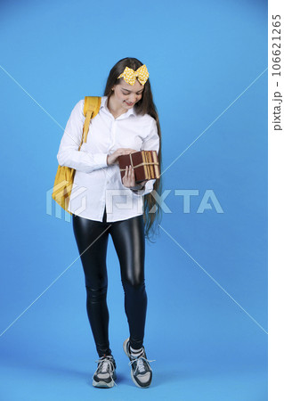 Happy, excited female student with yellow backpack and bow, looking at big bunch of books with pleasure, while standing in studio, isolated on blue background. Concept of studying. Happy, excited female student with yellow backpack and bow, looking at big bunch of books with pleasure, while standing in studio, isolated on blue background. Concept of studying. 106621265