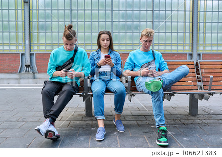 Group of teenage friends using smartphones sitting on chairs on train station platform Group of teenage friends using smartphones sitting on chairs on train station platform 106621383