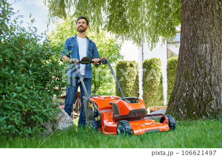 Handsome bearded man in blue shirt trimming lawn with electric mower on backyard. Low angle view of smiling male gardener standing with lawn trimmer, working in garden at day. Concept of gardening. Handsome bearded man in blue shirt trimming lawn with electric mower on backyard. Low angle view of smiling male gardener standing with lawn trimmer, working in garden at day. Concept of gardening. 106621497