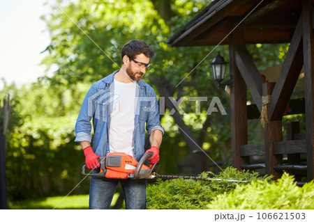 Professional male gardener in protective glasses shaping dense hedges with electric loppers on backyard. Front view of guy in shirt pruning shrub with trimmer at sunny day. Concept of hobby, work. Professional male gardener in protective glasses shaping dense hedges with electric loppers on backyard. Front view of guy in shirt pruning shrub with trimmer at sunny day. Concept of hobby, work. 106621503
