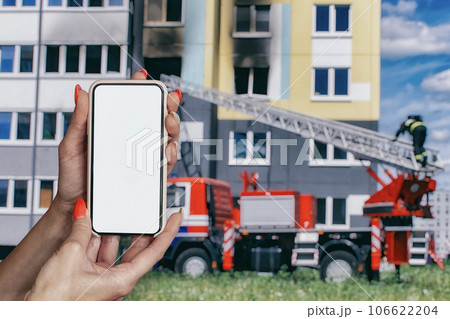 Girl holds a smartphone mockup with a white screen in her hands against the background of a fire engine with a firefighter and a burning house. Girl holds a smartphone mockup with a white screen in her hands against the background of a fire engine with a firefighter and a burning house. 106622204