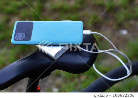 A portable charger charges a smartphone on the background of a bicycle and nature. Power Bank on a journey to charge gadgets. 106622206