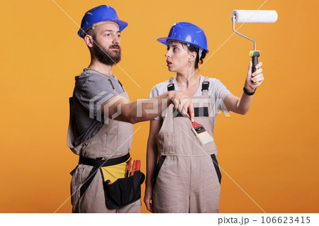 Young construction workers posing with painting tools, standing over yellow background. Team of constructors holding paintbrush and roller brush to work on refurbishment project in studio. Young construction workers posing with painting tools, standing over yellow background. Team of constructors holding paintbrush and roller brush to work on refurbishment project in studio. 106623415