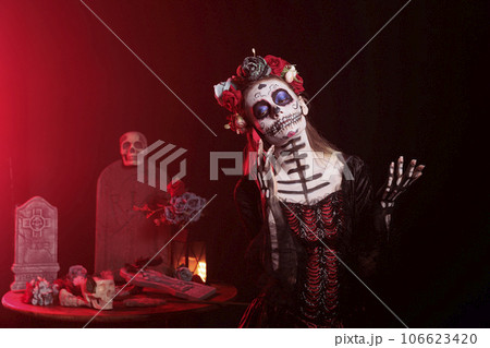 Scary woman talking on mobile phone call, wearing halloween make up and flowers crown in studio. La cavalera catrina using smartphone line on dios de los muertos mexican tradition. Scary woman talking on mobile phone call, wearing halloween make up and flowers crown in studio. La cavalera catrina using smartphone line on dios de los muertos mexican tradition. 106623420
