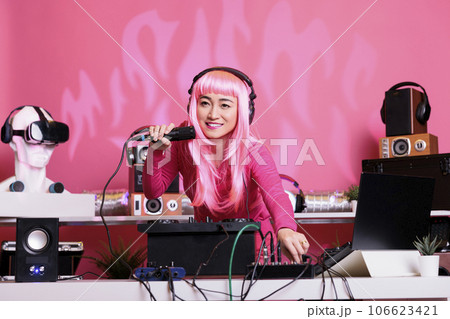 Artist with pink hair mixing techno song with electronic using professional turntables while enjoying talking with fans, having fun together during night time in club. Woman enjoying performing music 106623421