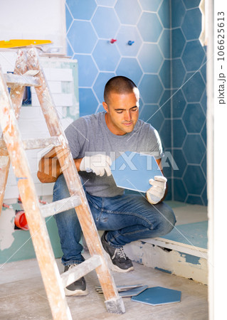 Portrait of concentrated young man sitting squat and making renovation repair. Tiler installing tile on wall Portrait of concentrated young man sitting squat and making renovation repair. Tiler installing tile on wall 106625613