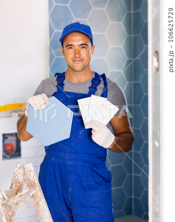 Skilled builder in work clothes posing with ceramic tiles and construction tools while repair and decoration in apartment 106625729
