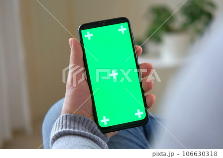 Young man sitting at home holding smartphone green mock-up screen in hand. Male person using chroma key mobile phone. Vertical mode. Touching, swiping display, tapping, surfing Internet social media Young man sitting at home holding smartphone green mock-up screen in hand. Male person using chroma key mobile phone. Vertical mode. Touching, swiping display, tapping, surfing Internet social media 106630318