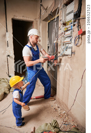 Male worker in safety helmet drilling wall with electric drill while little boy holding tool wire. Man and kid in work overalls installing electrical switch panel in apartment under renovation. Male worker in safety helmet drilling wall with electric drill while little boy holding tool wire. Man and kid in work overalls installing electrical switch panel in apartment under renovation. 106632523