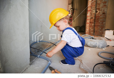 Little boy construction worker holding electrical wires in apartment under renovation. Child wearing work overalls and safety helmet while carrying cables. Little boy construction worker holding electrical wires in apartment under renovation. Child wearing work overalls and safety helmet while carrying cables. 106632532