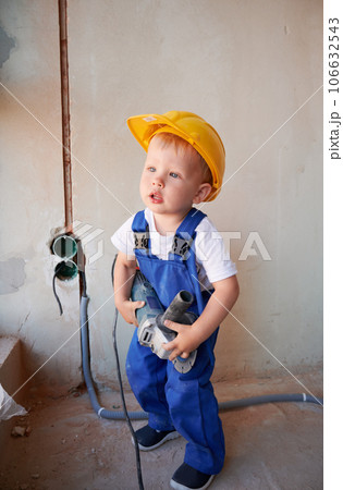 Full length of adorable little boy construction worker holding electric power grinding machine. Cute child wearing work overalls and safety helmet while standing in apartment under renovation. 106632543