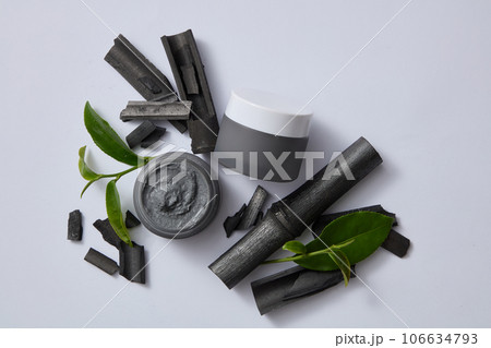Two mask jars with bamboo charcoal and green tea leaves on gray background. Mockup for advertising. Bamboo charcoal and tea are a duo of ingredients that help clean the skin and naturally moisturize Two mask jars with bamboo charcoal and green tea leaves on gray background. Mockup for advertising. Bamboo charcoal and tea are a duo of ingredients that help clean the skin and naturally moisturize 106634793