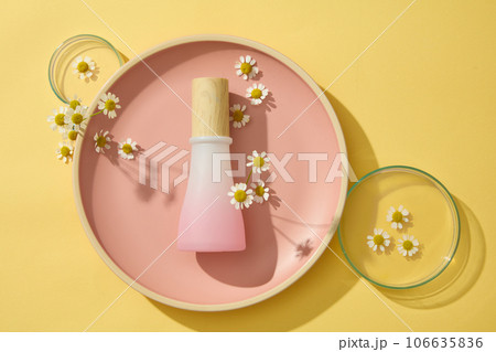 A pink glass bottle unbranded displayed on a round dishes with fresh chamomilla flowers and petri dishes on a yellow background. Herbs are widely used in cosmetics 106635836