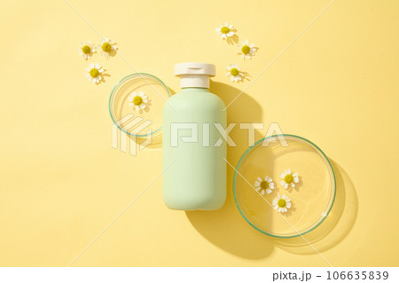 Top view of green plastic bottles of cream or lotion and with chamomilla flowers displayed on yellow background with petri dish. Blank label for design. Mockup scene for advertising 106635839