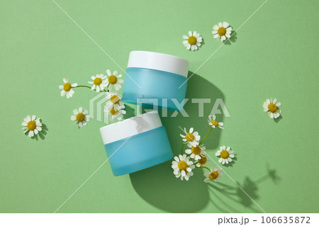 Concept for advertising cosmetics with natural ingredient - chamomilla. Two blue cream jars with flowers displayed on a green background. Chamomilla helps promote cell renewal for healthy glowing skin Concept for advertising cosmetics with natural ingredient - chamomilla. Two blue cream jars with flowers displayed on a green background. Chamomilla helps promote cell renewal for healthy glowing skin 106635872
