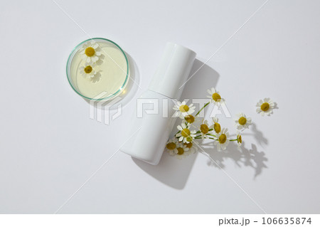 A white cosmetic bottle without label decorated with fresh flowers and petri dish containing essence of chamomilla on a white background. Minimal scene for advertising and branding cosmetic A white cosmetic bottle without label decorated with fresh flowers and petri dish containing essence of chamomilla on a white background. Minimal scene for advertising and branding cosmetic 106635874