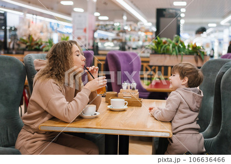 Young curly haired mother and her little son are having breakfast or lunch in a cafe, sitting at a table. Family weekend 106636466