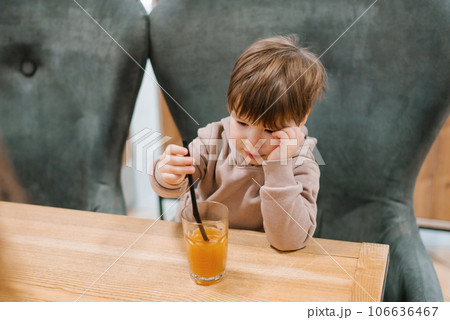 Four year old boy sits at a table in a cafe and looks at a glass of orange juice, holding a straw in his hands 106636467