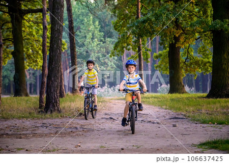 Two little kid boys in colorful casual clothes in summer forest park driving bicycle. Active children cycling on sunny fall day in nature. Safety, sports, leisure with kids concept Two little kid boys in colorful casual clothes in summer forest park driving bicycle. Active children cycling on sunny fall day in nature. Safety, sports, leisure with kids concept 106637425