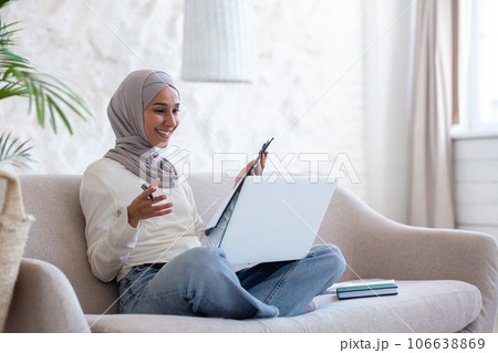 Young Muslim businesswoman in hijab working remotely, sitting on sofa at home in lotus position, holding documents in hands and smiling talking via laptop video call. 106638869