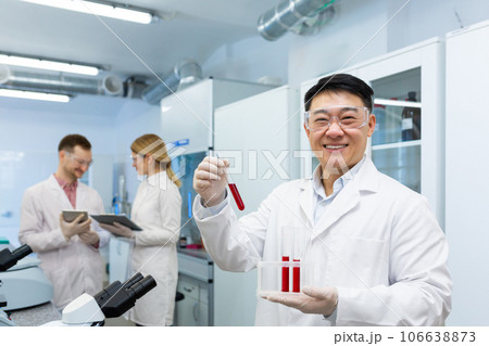Portrait of a smiling young doctor of Asian descent standing in the laboratory and holding flasks with blood samples. Colleagues are discussing work behind. Portrait of a smiling young doctor of Asian descent standing in the laboratory and holding flasks with blood samples. Colleagues are discussing work behind. 106638873