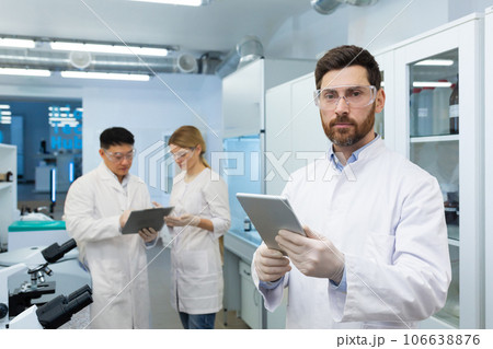 Portrait of a young male scientist, a doctor standing in a research medical laboratory and holding a tablet in his hands, looking at the camera. Behind him, colleagues discussing the results. 106638876