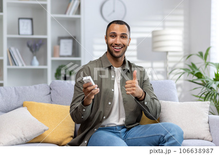 Portrait of a young African American man sitting on the sofa at home and holding the air conditioner remote control, smiling at the camera and showing the super gesture with his finger. 106638987