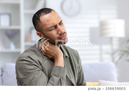Tired young African American man sitting at home and holding his neck with eyes closed, suffering from pain and overtiredness. Close-up photo. Tired young African American man sitting at home and holding his neck with eyes closed, suffering from pain and overtiredness. Close-up photo. 106639003