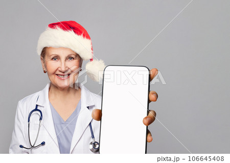 Christmas portrait of smiling woman doctor in Santa hat showing smartphone with blank emptydisplay Christmas portrait of smiling woman doctor in Santa hat showing smartphone with blank emptydisplay 106645408