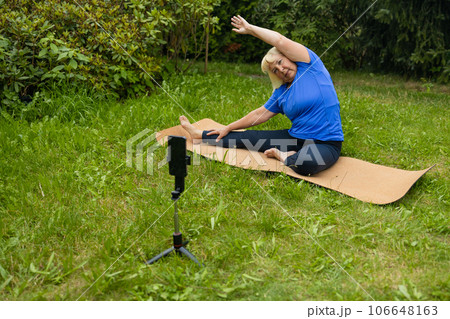 Fit mature 50s female in casual clothes exercising outdoors, practicing yoga on mat and talking at smartphone camera, making video content for followers, looking at webcam on mobile phone on tripod Fit mature 50s female in casual clothes exercising outdoors, practicing yoga on mat and talking at smartphone camera, making video content for followers, looking at webcam on mobile phone on tripod 106648163