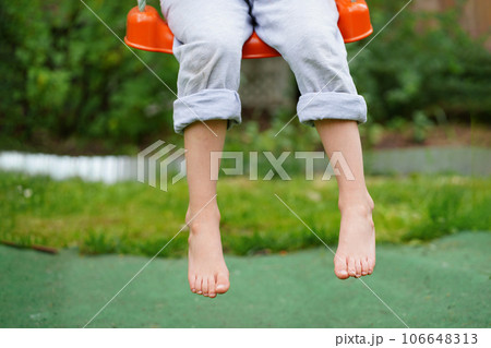 Children's legs hanging from the swing. Closeup of happy kid sitting and swinging, sunny summer day, countryside background. Child feet barefoot Children's legs hanging from the swing. Closeup of happy kid sitting and swinging, sunny summer day, countryside background. Child feet barefoot 106648313