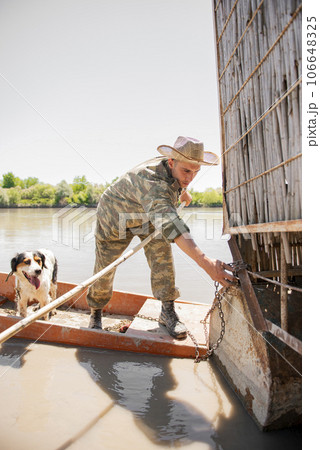 Young fisherman in panama hat untying rowing boat from wharf, while going fishing with dog outside. Front view of concentrated man in camo untangling metal chain of skiff at dock. Concept of fishery. Young fisherman in panama hat untying rowing boat from wharf, while going fishing with dog outside. Front view of concentrated man in camo untangling metal chain of skiff at dock. Concept of fishery. 106648325