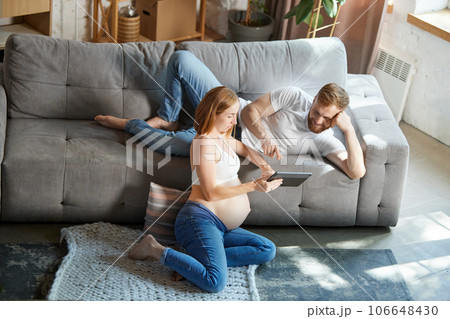 Young couple, future parents sitting at home and looking on tablet. Expecting baby, learning information Young couple, future parents sitting at home and looking on tablet. Expecting baby, learning information 106648430