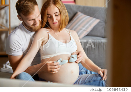 Care and love. Happy man and pregnant woman, sitting on couch, holding little shoes on belly. Parents expecting baby 106648439
