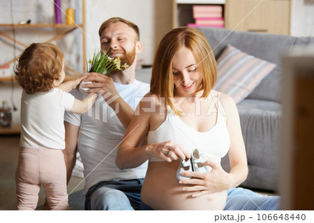 Happy man playign with daughter and pregnant woman holding little shoes on her belly. Expecting baby Happy man playign with daughter and pregnant woman holding little shoes on her belly. Expecting baby 106648440