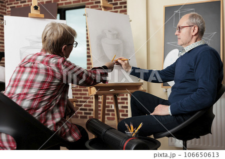 Retired couple talking discussing artwork while learning new skill. Senior man and woman sitting at easels learn to draw, attending group art class together. Creative activities to do with family 106650613