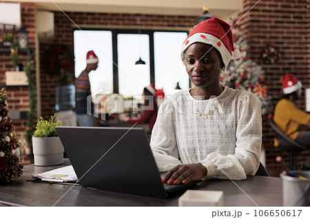 African american woman company manager wearing santa claus hat typing on laptop in christmas decorated office. Employee sitting at desk and working on computer in festive workspace African american woman company manager wearing santa claus hat typing on laptop in christmas decorated office. Employee sitting at desk and working on computer in festive workspace 106650617
