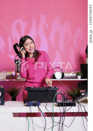 Cheerful performer with headphones playing electronic song at mixer console, standing at dj table having fun with fans at night in club. Artist performing techno music in studio over pink background 106650643