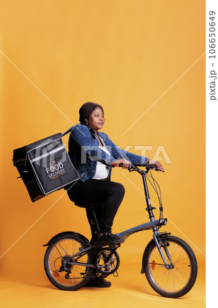 Pizzeria worker carrying food takeaway backpack standing on bike in studio with yellow background, delivering pizza order for dinner. Food transportation service and takeaway concept Pizzeria worker carrying food takeaway backpack standing on bike in studio with yellow background, delivering pizza order for dinner. Food transportation service and takeaway concept 106650649