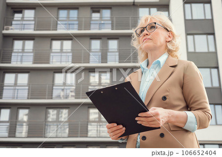 Senior female real estate agent stands holding clipboard in hands against residential complex in daytime Senior female real estate agent stands holding clipboard in hands against residential complex in daytime 106652404