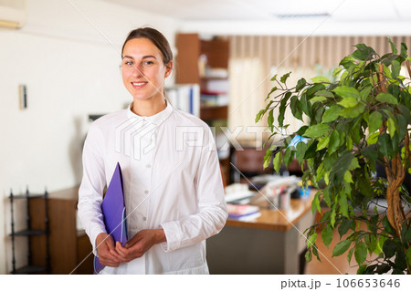 Portrait of a young female manager standing in the office with a folder of documents Portrait of a young female manager standing in the office with a folder of documents 106653646